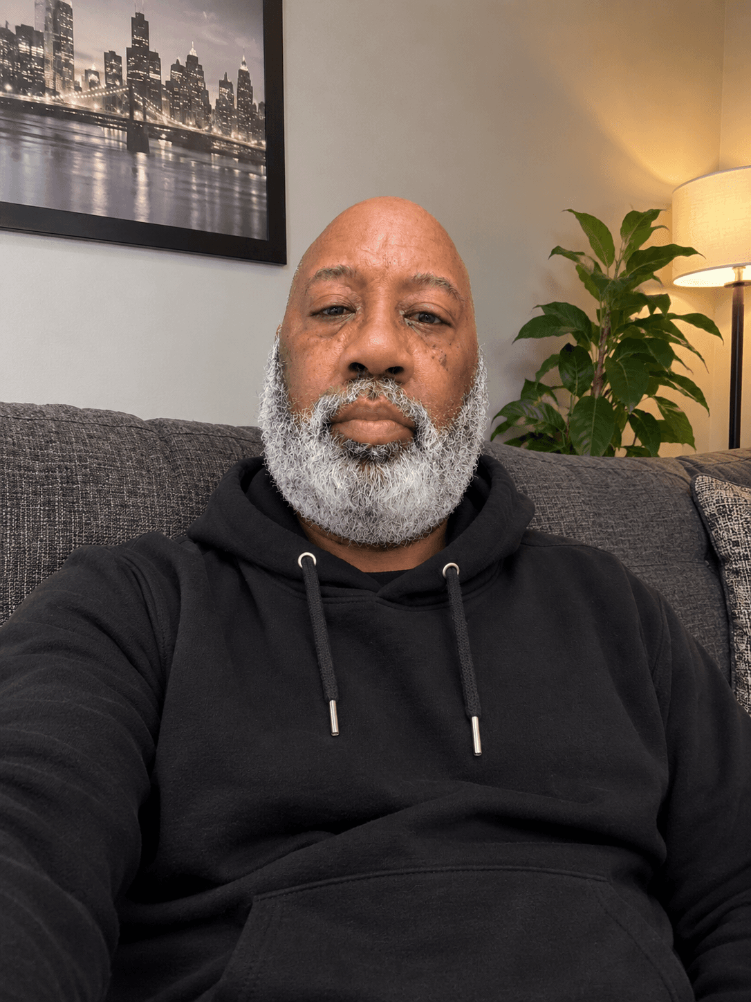 Bald Black man with a white beard wearing a black hoodie on a grey couch.